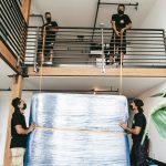 Four movers carefully lift a large wrapped object using straps indoors.